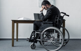 Depressed disabled businessman sitting in wheelchair at workplace with bowed head