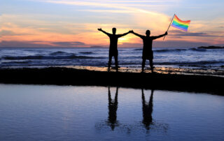 Silhouette of a gay couple holding a rainbow pride flag at sunset.